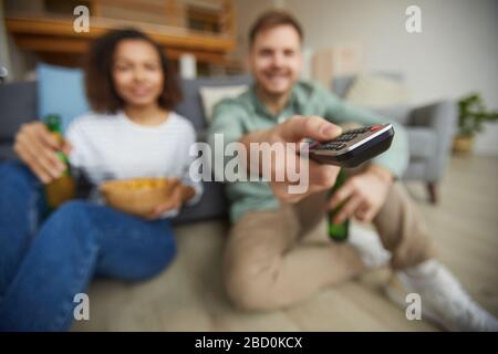 Blurred portrait of young mixed-race couple watching TV at home and holding remote while sitting on floor in cozy apartment, focus on foreground Stock Photo