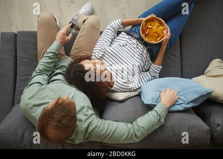 Above view portrait of young mixed-race couple watching TV at home and eating snacks while lying on comfortable sofa, copy space Stock Photo