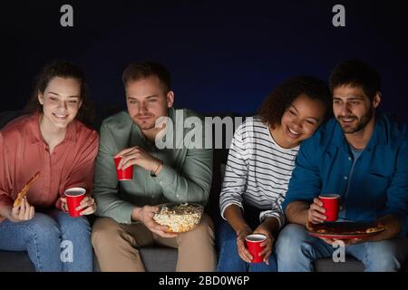 Portrait of two young couples watching movies at home while eating snacks and popcorn sitting on sofa in dark room, copy space above Stock Photo