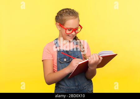 Early child education. Portrait of creative smart little girl in bright red glasses writing in notebook, drawing with pencils, taking notes, school ch Stock Photo