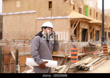 Foreman talking by smartphone and holding drawing plan at construction site. Stock Photo