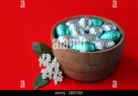 A woden bowl filled with silver and aqua green foil covered easter eggs. In close up against a bright orange red background with white spring flower. Stock Photo