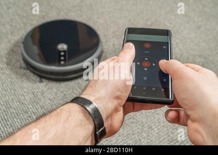 Male hands are holding the phone and through wifi controls the robot vacuum cleaner Stock Photo