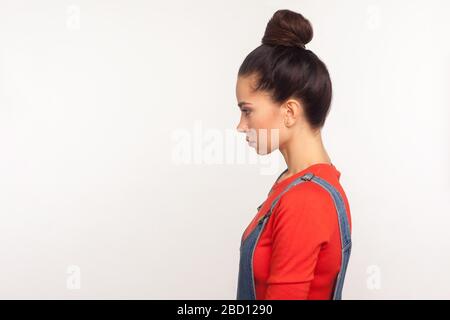 Profile of upset depressed girl with hair bun in denim overalls standing with head down and unhappy look, expressing sadness, worrying about problems.