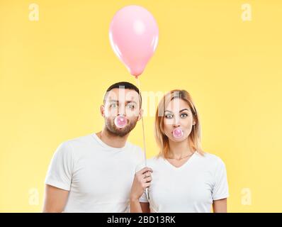 Young couple blowing bubble with chewing gum and holding pink air balloon on yellow background Stock Photo