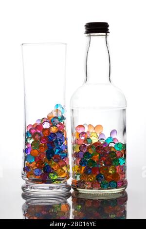 A bottle and glass filled with multicolored gel balls. Reflection on a glossy surface. Bright background.