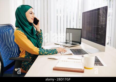 Smiling young muslim software developer talking on phone when checking programming code on computer screen Stock Photo