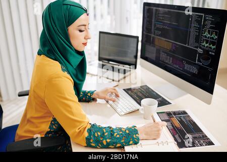 young female software developer using desktop computer while writing ...