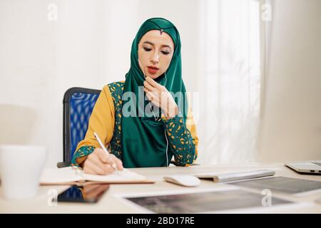 Young beautiful muslim businesswoman in traditional dress talking on phone with colleague and taking notes in planner or journal Stock Photo