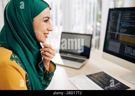 Cheerful smiling muslim young woman wearing earphones, programming on computer and talking to coworker Stock Photo