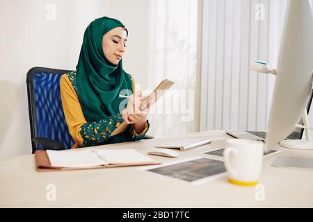 Serious muslim female entrepreneur sitting at her desk and taking notes in planner Stock Photo