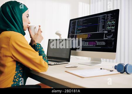 Pensive female muslim coder drinking tea and looking at programming code on computer screen Stock Photo