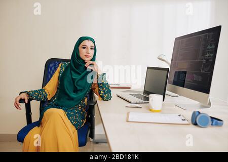 Confident muslim female software developer in traditional dress in hijab sitting at her office table Stock Photo