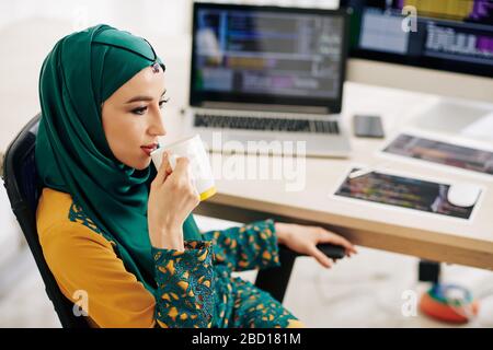 Pensive pretty young muslim woman sitting at desk and drinking big mug of tea or coffee Stock Photo