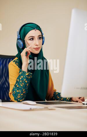 Pensive muslim female programmer in traditional dress looking at screen of computer Stock Photo
