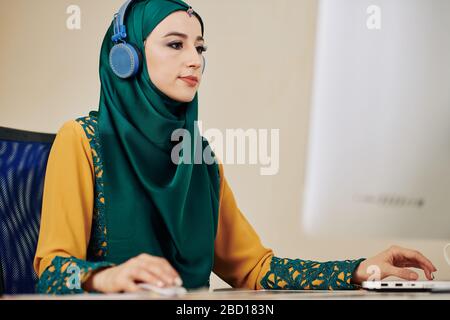 Young serious muslim woman in hijab listening to music in headphones when working on computer Stock Photo