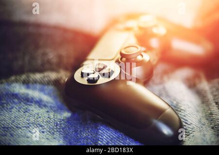 A modern black gamepad for playing video games lies on a plaid blanket on the bed, illuminated by a bright light. Home entertainment. Stock Photo