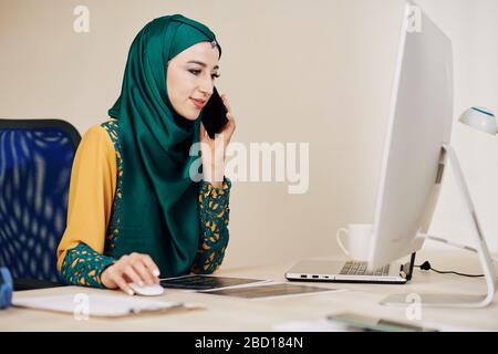 Smiling muslim female software developer talking on phone with client Stock Photo