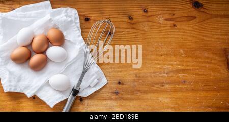 Top view of three white and four brown eggs on a clean white cloth next to a whisker next to empty space on a wooden background. Stock Photo