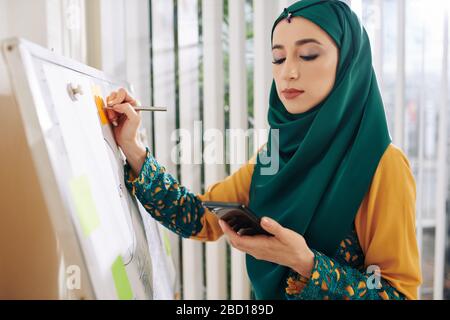 Young pretty muslim businesswoman in traditional clothes checking smartphone when writing ideas on colorful stickers Stock Photo