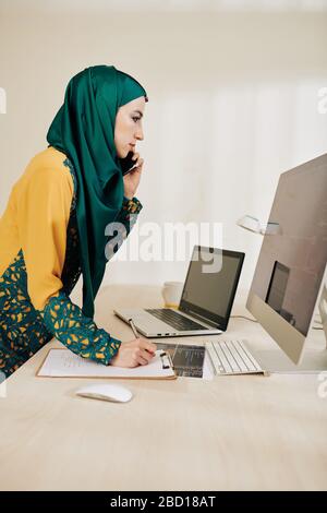 Frowning serious muslim businesswoman reading article on computer screen and talking on phone with client Stock Photo
