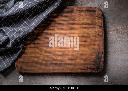 Top view of wooden dark brown vintage chopping board on dark metalic background with black checked cloth partial covering. Stock Photo