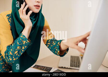 Cropped image of muslim business lady in traditional clothes pointing at computer screen and talking on phone with colleague Stock Photo