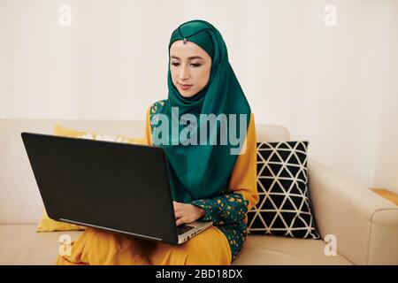 Young muslim female freelancer sitting on sofa at home and working on laptop Stock Photo