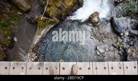 A point of view image of a hiker looking at their hiking boots and peering over the edge of a wooden bridge with no railing over a mountain stream Stock Photo