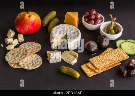 Cheeseboard with camembert, cheddar, crackers, olives, figs, dates and pickles on black texture background - focus on front Stock Photo