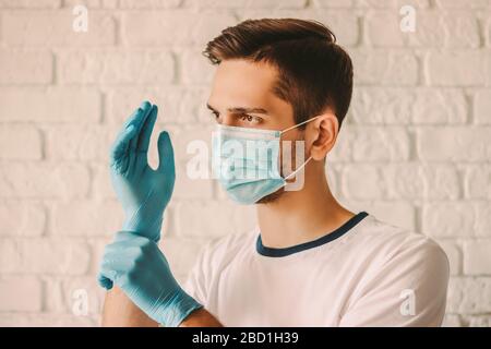 Young man hospital staff in medical mask on face wearing blue latex gloves on hands. Professional doctor surgeon in protective face mask putting on me Stock Photo