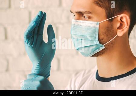 Professional medic in protective mask on face putting on blue latex gloves on hands. Young man surgeon in face mask wearing sterile medical glove. Med Stock Photo