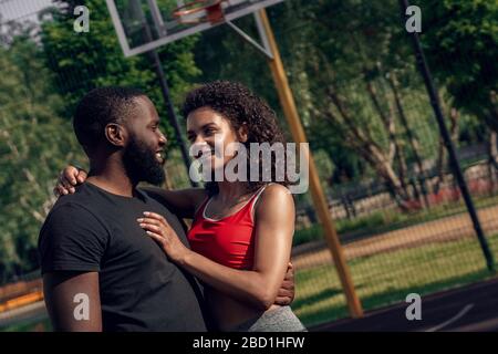 Outdoors Activity. African couple standing on basketball court hugging looking at each other smiling in love close-up Stock Photo