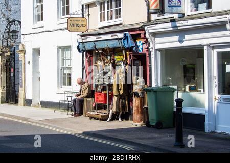 A man sat in the sun outside an antiques and curiousity  shop with various items displayed on the pavement Stock Photo