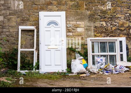 An old PVC door and some wooden window frames leant against a wall with assorted rubbish and weeks Stock Photo