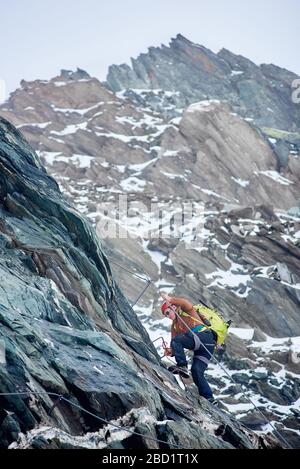 Mountaineer with backpack using fixed rope to climb high rocky mountain. Man climber ascending alpine ridge and trying to reach mountaintop. Concept of mountaineering and alpine rock climbing. Stock Photo