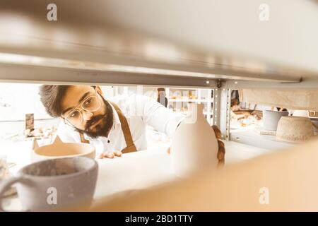 Young attractive master potter pulls finished products from the shelves for inspection Stock Photo