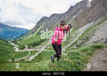 Side view of female hiker with trekking poles walking uphill in mountain valley. Woman in sport jacket looking down, smiling while hiking alone in mountains. Concept of travelling, hiking and tourism. Stock Photo