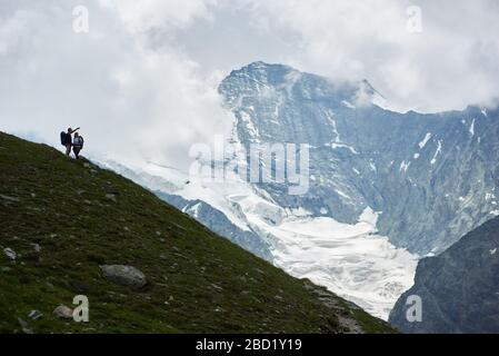 Silhouette of loving couple with backpacks looking at mountains scenery, man pointing with hand. Trekking, tourists reaching peak together. Wild nature with amazing views. Sport tourism in Alps. Stock Photo
