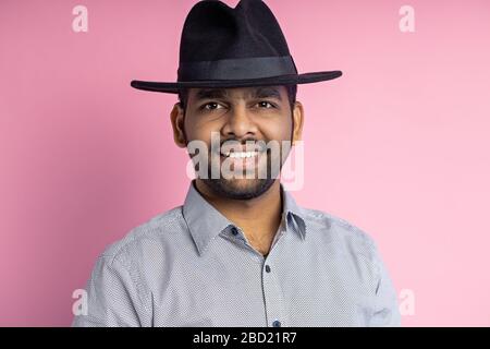 Closeup shot of pleased joyful young indian male with stubble, wearing casual shirt, black hat, looking with smile and pointing to side with forefinge Stock Photo