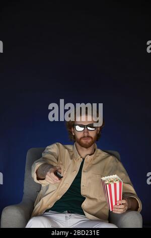 Vertical portrait of modern bearded man watching movie and wearing stereo glasses while sitting on couch in dark room, copy space Stock Photo