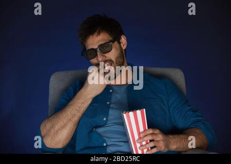 Portrait of bearded man wearing 3D glasses eating popcorn while watching movie at home alone, copy space Stock Photo