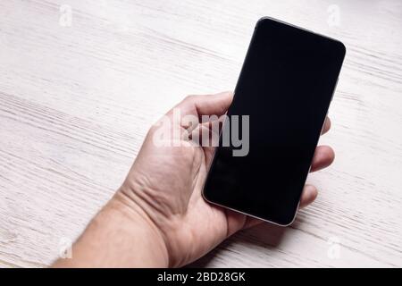 hand holding a smart phone with blank screen on white wooden table, technology and communication concept, copy space for text, selective focus Stock Photo