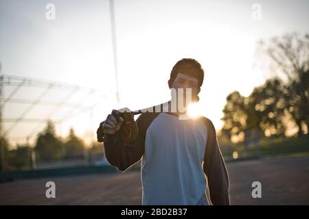 Portrait of a teenage boy carrying a baseball mitt and baseball bat over his shoulder in the sunlight. Stock Photo