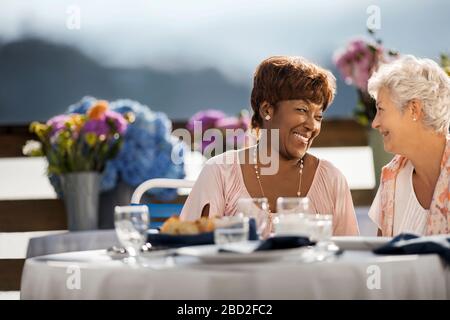 Two mature female friends smile happily at each other as they enjoy a meal at an outdoor restaurant table. Stock Photo