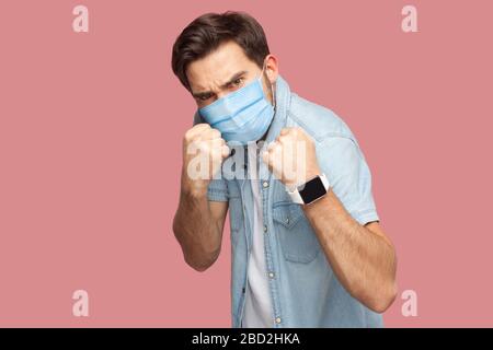Portrait of angry man with surgical medical mask in blue casual style shirt standing with boxing fists and looking at camera and ready to attack. indo Stock Photo