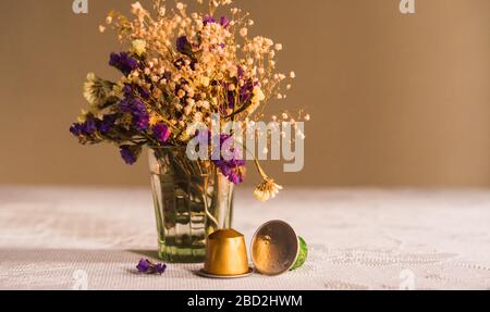 Two Nespresso coffee capsules on the table in front of the purple and white flowers in a glass vase on a white textile in a dark background Stock Photo