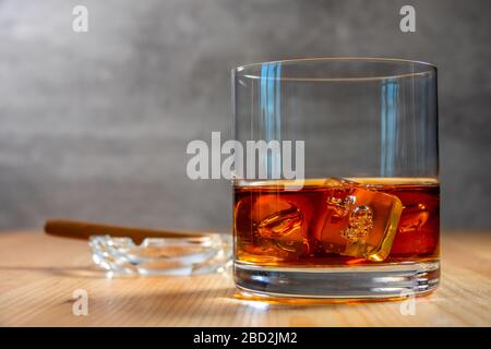 On the table is a glass of whiskey with ice cubes. An ashtray with a cigar in the background in defocus Stock Photo