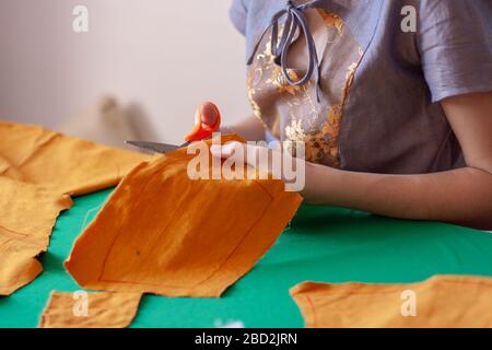 Girl seamstress cuts the fabric with scissors. Pattern for face masks. Orange fabric. Focus on scissors and fabric. Horizontal. Stock Photo