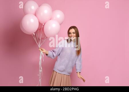 Smiling carefree girl in knitted sweater dancing with pastel pink air balloons isolated on pink background. Beautiful happy young woman on a birthday holiday. space for text Stock Photo
